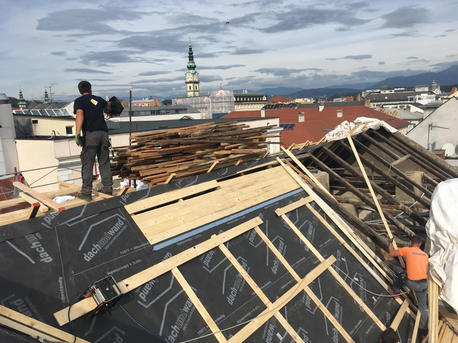 Zwei Arbeiter auf einem offenen Dachstuhl über den Dächern von Klagenfurt